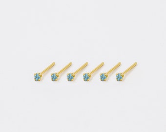 May include: Five gold nose rings with a small blue gemstone on each. The rings are arranged in a row on a white background.