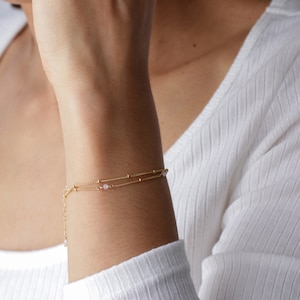 Puede incluir: Un delicado brazalete de cadena dorada con dos hebras y peque&ntilde;as cuentas rosas.