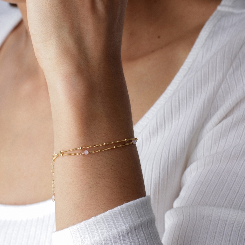 Puede incluir: Un delicado brazalete de cadena dorada con dos hebras y peque&ntilde;as cuentas rosas.