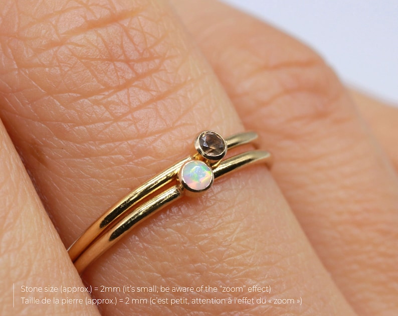 May include: Two gold rings with small gemstones. The top ring has a light brown gemstone and the bottom ring has a white opal gemstone. The rings are on a finger.