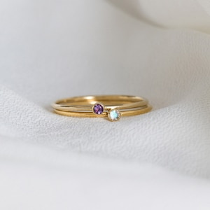 May include: Two gold rings with small gemstones. The top ring has a light blue gemstone and the bottom ring has a purple gemstone.