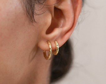 Puede incluir: Un par de pendientes de aro dorados martillados. Los pendientes son pequeños y delicados, y se llevan en el lóbulo de la oreja.