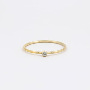 Può includere: Un delicato anello d'oro con una piccola gemma azzurra al centro. L'anello ha una fascia sottile e un design semplice ed elegante. La gemma di forma ovale sembra essere saldamente incastonata nella fascia d'oro.