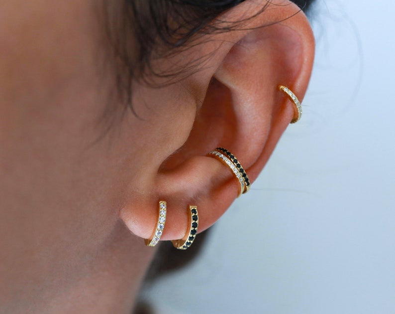 May include: A close-up of a person's ear with multiple gold hoop earrings. The earrings are adorned with small black and white stones.