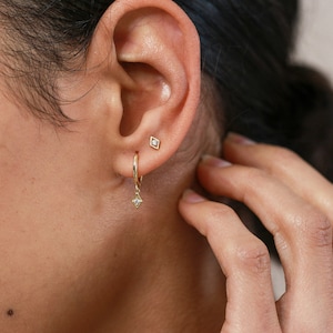 May include: A pair of gold earrings with small diamond accents. The earrings are a hoop style with a dangling diamond charm. The earrings are worn in the ear of a person with dark hair.