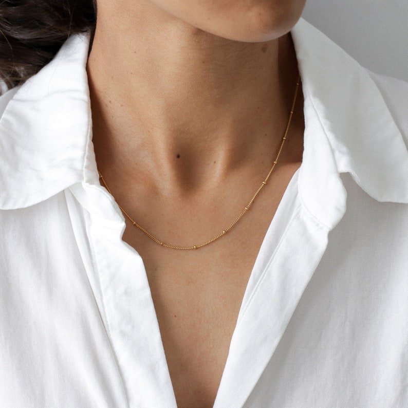 May include: A delicate gold chain necklace with small beads. The necklace is worn by a person with a white shirt.