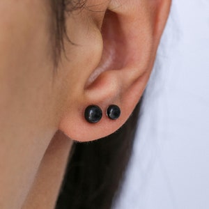 Könnte beinhalten: Ein Paar schwarze Ohrstecker. Die Ohrringe sind klein und rund und werden in den Ohrläppchen getragen.