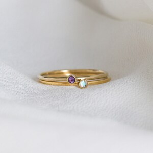 May include: Two gold rings with small gemstones. The top ring has a light blue gemstone and the bottom ring has a purple gemstone.