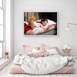 May include: A framed print shows a woman wearing a sleep mask, lying on a bed with a ginger cat. The cat is sitting on the bed. The bed has white pillows and sheets. The room has a white wall and a window.