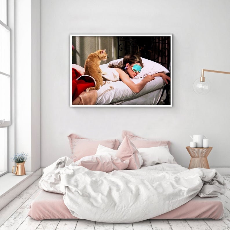 May include: A framed print shows a woman wearing a sleep mask, lying on a bed with a ginger cat. The cat is sitting on the bed. The bed has white pillows and sheets. The room has a white wall and a window.