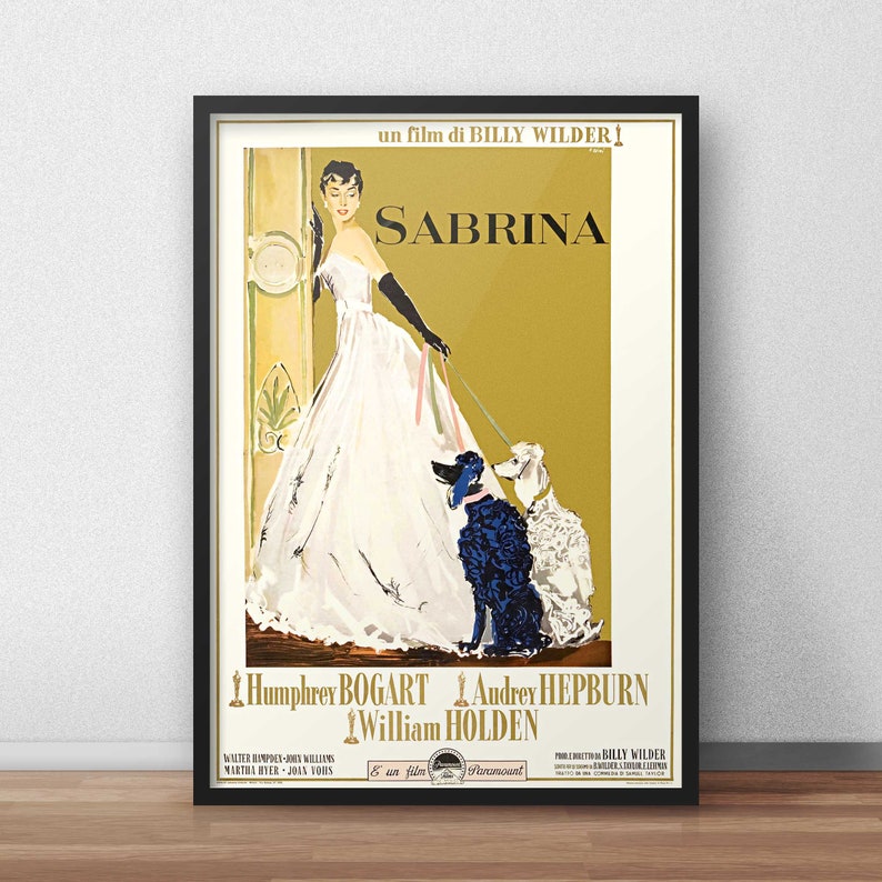 May include: A framed movie poster for Sabrina, a film by Billy Wilder. The poster features an illustration of a person in a white gown and black gloves holding leashes for two poodles, one black and one white, against a gold background.