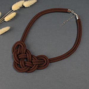 Può includere: Una collana in corda marrone con un elemento centrale annodato. La collana presenta un design a doppio filo e una chiusura color argento. La sezione annodata è una caratteristica importante, che aggiunge una texture unica al gioiello. La collana è esposta su una superficie grigia.