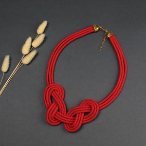 May include: A red rope necklace with a knotted design. The necklace features three parallel strands that form a circular shape, with two intricate knots at the center. Gold-colored metal accents are at the clasp.