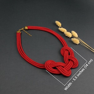 Pode incluir: Um colar de corda vermelha com um design de n&oacute;s, com cerca de 14 cm de largura. O colar apresenta um fecho dourado e &eacute; exibido sobre um fundo escuro. Flores secas no fundo.