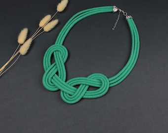 Puede incluir: Un collar verde vibrante con un diseño anudado. El collar está hecho de un material grueso y tejido y presenta un cierre plateado. El diseño incluye un nudo central y tres hebras paralelas. El fondo es gris oscuro.