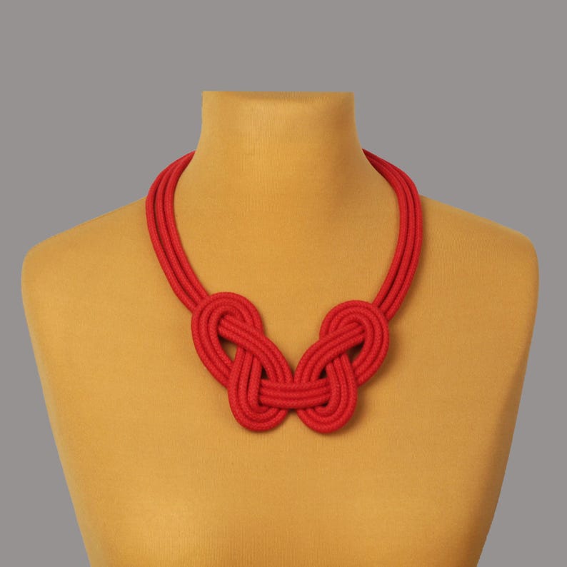 Pode incluir: Um colar vermelho vibrante com um design de n&oacute; &uacute;nico. O colar &eacute; exibido em um manequim, mostrando sua cor ousada e sua intrincada textura semelhante a uma corda. O design apresenta dois n&oacute;s grandes e sim&eacute;tricos conectados por uma dupla mecha.