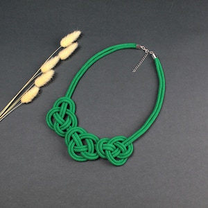 Puede incluir: Un collar verde brillante con tres secciones anudadas. El collar está hecho de un material grueso y tejido y tiene un cierre plateado. Se muestra sobre una superficie gris oscuro con flores secas de tono amarillo.