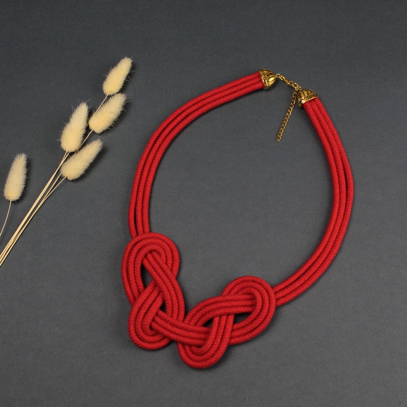 May include: A red rope necklace with a knotted design. The necklace features three parallel strands that form a circular shape, with two intricate knots at the center. Gold-colored metal accents are at the clasp.