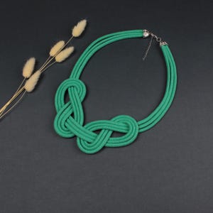 Pode incluir: Um colar de corda verde com um design de n&oacute;s. O colar &eacute; feito de tr&ecirc;s fios de corda e tem um fecho prateado. O colar &eacute; exibido em uma superf&iacute;cie cinza escuro, com flores secas ao fundo.