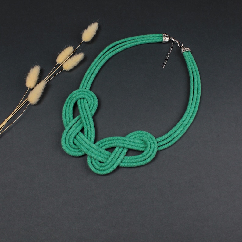Pode incluir: Um colar de corda verde com um design de n&oacute;s. O colar &eacute; feito de tr&ecirc;s fios de corda e tem um fecho prateado. O colar &eacute; exibido em uma superf&iacute;cie cinza escuro, com flores secas ao fundo.