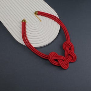 Pode incluir: Um colar de corda vermelha com um design de n&oacute;s. O colar tem um fecho e extremidades douradas. O colar &eacute; exibido em uma superf&iacute;cie branca e curva sobre um fundo cinza escuro.
