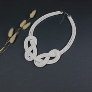 Pode incluir: Um colar de corda branca com um design de n&oacute;s. O colar apresenta uma s&eacute;rie de n&oacute;s entrela&ccedil;ados e um fecho prateado. O colar &eacute; exibido sobre uma superf&iacute;cie cinza escuro, com grama ornamental seca no fundo.