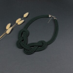 Pode incluir: Colar de corda verde escuro com um design de n&oacute;s. O colar apresenta um fecho prateado e &eacute; exibido sobre uma superf&iacute;cie cinza escuro. Um raminho de flores secas est&aacute; no fundo.