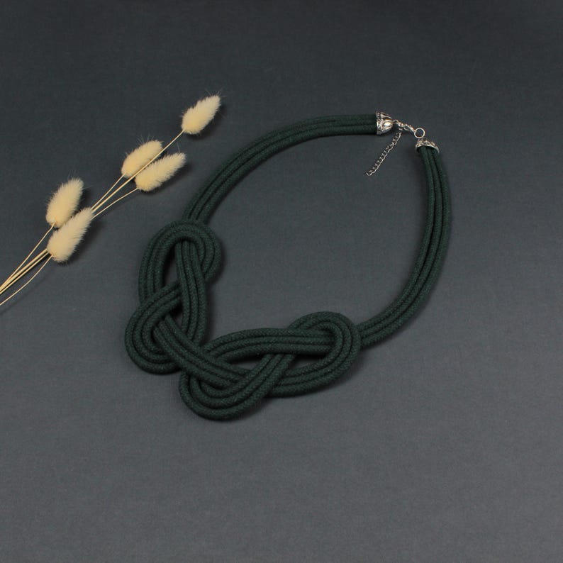 Pode incluir: Colar de corda verde escuro com um design de n&oacute;s. O colar apresenta um fecho prateado e &eacute; exibido sobre uma superf&iacute;cie cinza escuro. Um raminho de flores secas est&aacute; no fundo.