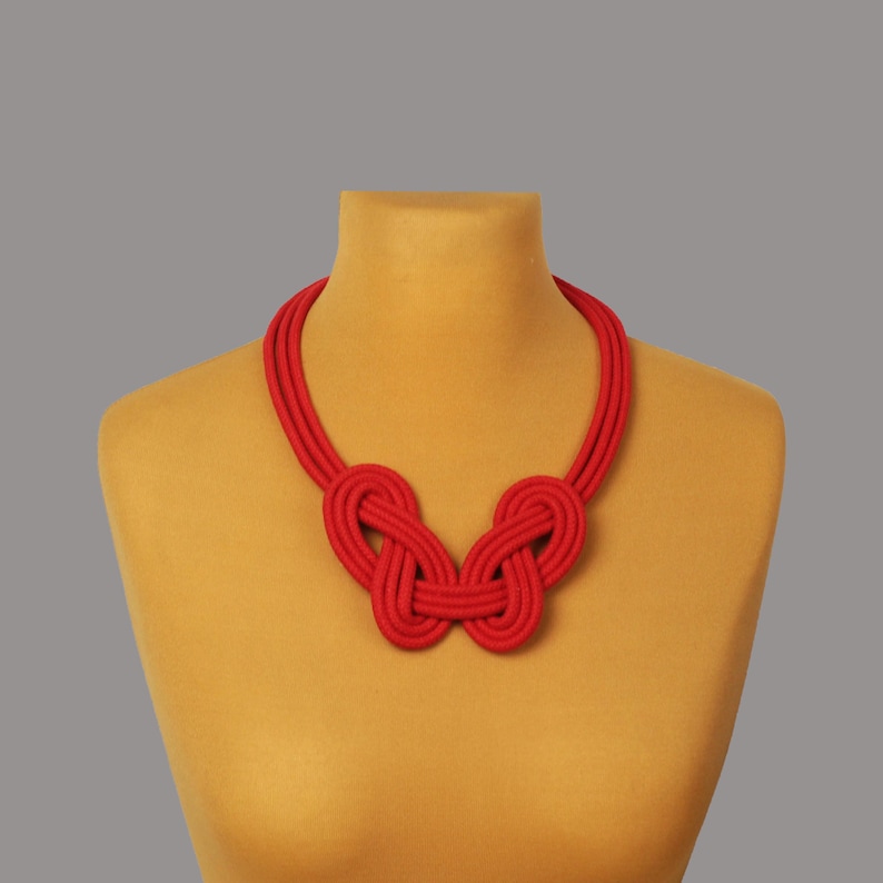 Pode incluir: Um colar vermelho vibrante exibido em um manequim. O colar apresenta um design com n&oacute;s no centro, com v&aacute;rios fios formando a base. A cor vermelha brilhante o torna um acess&oacute;rio marcante.