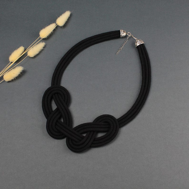 Pode incluir: Um colar de corda preta com um design de n&oacute;s. O colar apresenta uma s&eacute;rie de n&oacute;s no centro e um fecho prateado. O colar &eacute; exibido em uma superf&iacute;cie cinza, com um raminho de flores secas ao fundo.