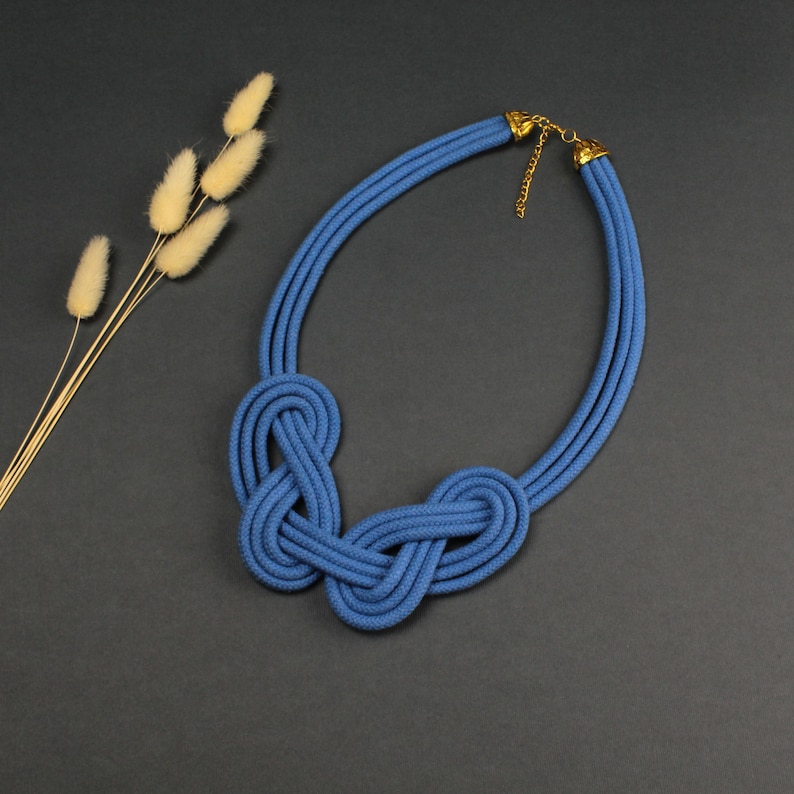Pode incluir: Um colar de corda azul com um design de n&oacute;s. O colar apresenta tr&ecirc;s fios paralelos que formam um la&ccedil;o, com um fecho e corrente dourados. A se&ccedil;&atilde;o de n&oacute;s est&aacute; no centro.