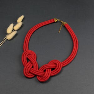 Pode incluir: Um colar arrojado de corda vermelha com um design de n&oacute;s. O colar apresenta tr&ecirc;s fios de corda vermelha que formam dois n&oacute;s grandes no centro. O colar tem um fecho dourado e &eacute; exibido em uma superf&iacute;cie cinza escuro.
