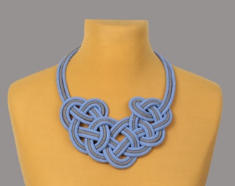 May include: A blue and gray rope necklace displayed on a mannequin. The necklace features a series of intertwined knots in a light blue and gray color scheme. The necklace is designed to be worn around the neck.