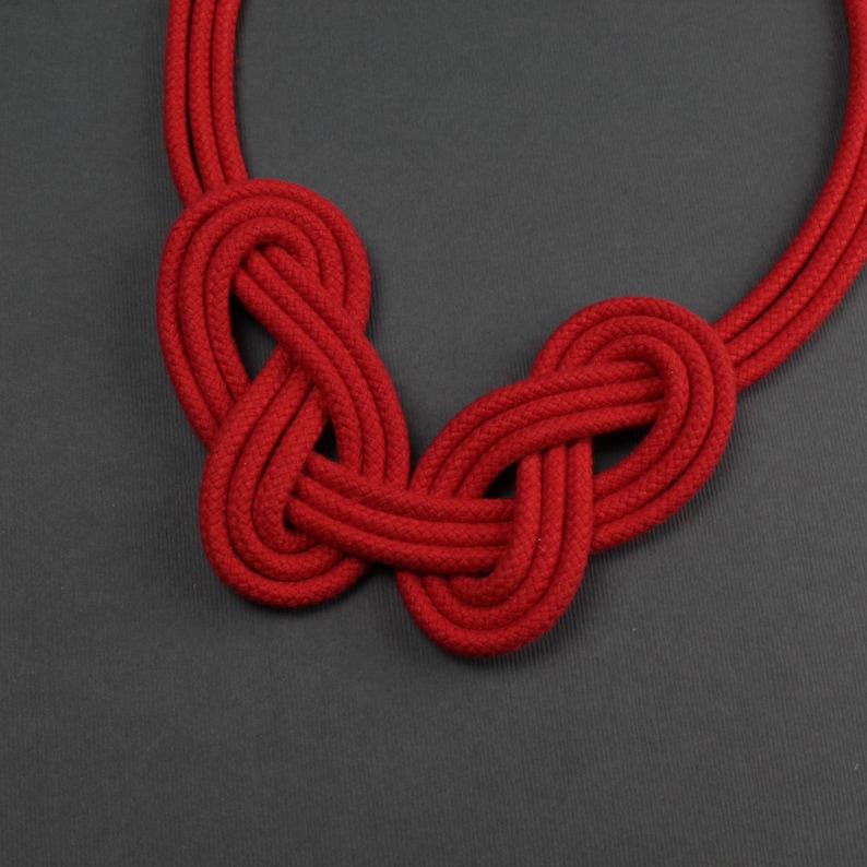 Pode incluir: Um colar de corda vermelha com dois intrincados desenhos de n&oacute;s. O colar &eacute; feito de corda vermelha grossa e tecida, com uma textura suave. Os n&oacute;s s&atilde;o o ponto focal, criando uma pe&ccedil;a ousada e atraente. O fundo &eacute; cinza s&oacute;lido.