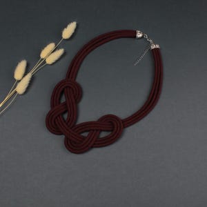 Pode incluir: Um colar de corda bord&ocirc; com um design de n&oacute;s. O colar apresenta um fecho prateado e uma s&eacute;rie de fios de corda bord&ocirc;. O colar &eacute; exibido em uma superf&iacute;cie cinza escuro, com flores secas ao fundo.