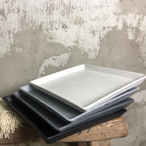 Könnte beinhalten: Ein Satz von vier quadratischen Tabletts in verschiedenen Grautönen, übereinander gestapelt. Die Tabletts bestehen aus einem strukturierten Material, möglicherweise Beton oder Keramik.