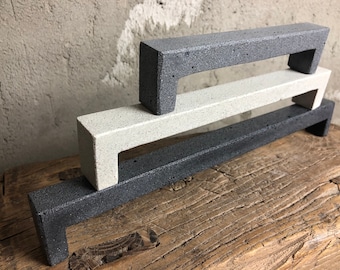 May include: Three concrete cabinet door pulls in different shades of gray. The pulls are stacked on top of each other, with the darkest gray pull on the bottom and the lightest gray pull on the top.