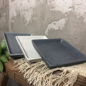 May include: Three square ceramic trays in different shades of gray. The trays are arranged in a row on a wooden surface with a fringe rug.