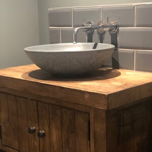 May include: A gray concrete vessel sink with a chrome faucet mounted on a gray tiled wall. The sink is set on a wooden vanity with a cabinet door.
