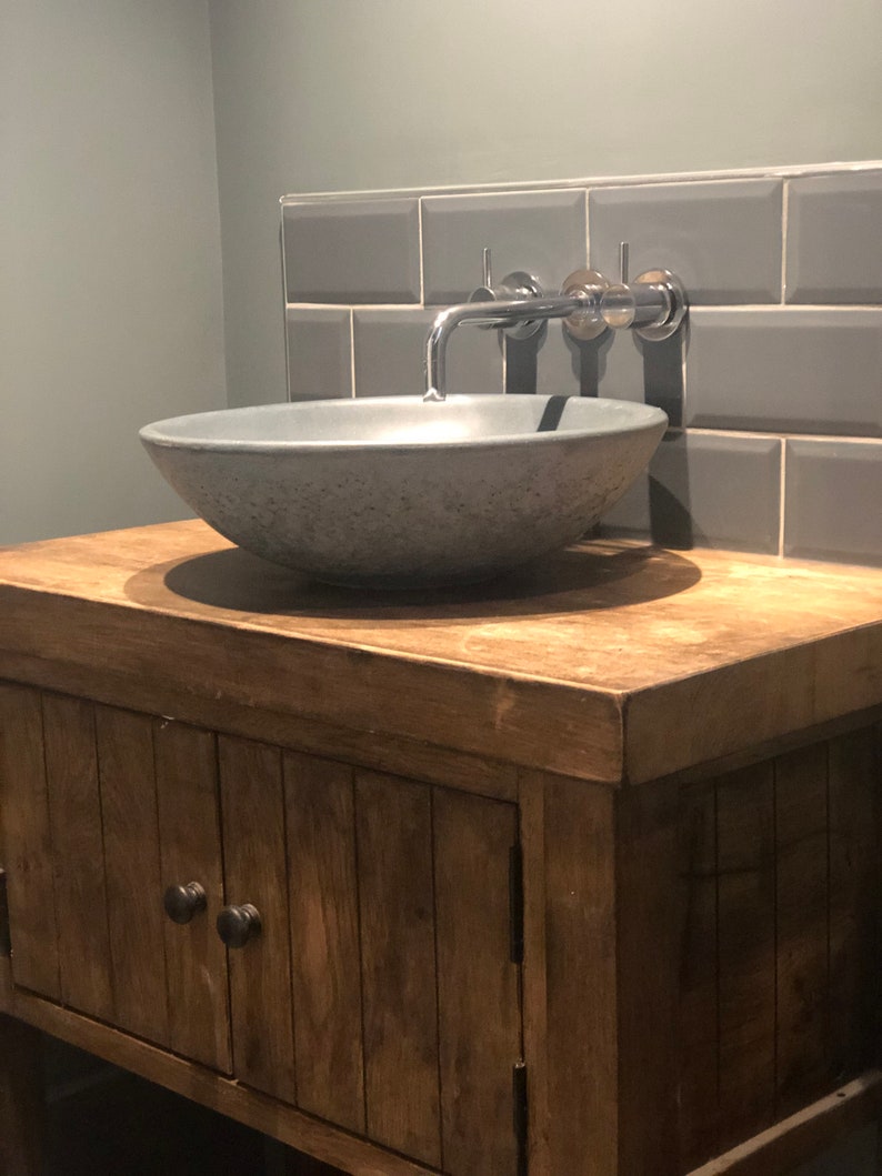 May include: A gray concrete vessel sink with a chrome faucet mounted on a gray tiled wall. The sink is set on a wooden vanity with a cabinet door.