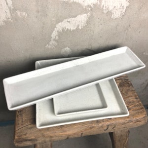 May include: Three white ceramic rectangular serving trays with a speckled finish. The trays are stacked on top of each other on a wooden surface.