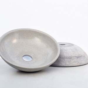 May include: Two gray ceramic bowls, one with a drain hole, are sitting on a gray surface. The bowls are both round and have a smooth, slightly textured surface.