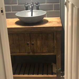 May include: A wooden bathroom vanity with a gray ceramic sink and a chrome faucet. The vanity has a cabinet with a wooden door and a lower shelf. The vanity is set against a tiled wall with gray subway tiles.