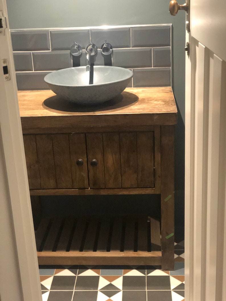 May include: A wooden bathroom vanity with a gray ceramic sink and a chrome faucet. The vanity has a cabinet with a wooden door and a lower shelf. The vanity is set against a tiled wall with gray subway tiles.
