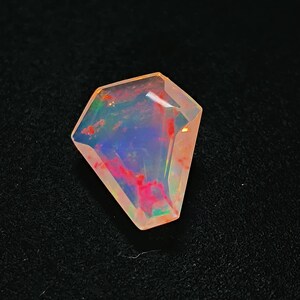 May include: A coffin-shaped faceted opal gemstone displaying a translucent body with vibrant flashes of red, blue, and green play-of-color throughout its surface. The stone features sharp, angular edges and a polished, reflective top surface.