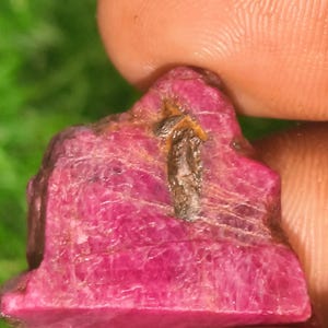 May include: A raw, unpolished ruby gemstone with a vibrant magenta-pink hue and a rough, crystalline texture. The stone features a prominent, dark, elongated inclusion running vertically across its surface, showing natural mineral variations.