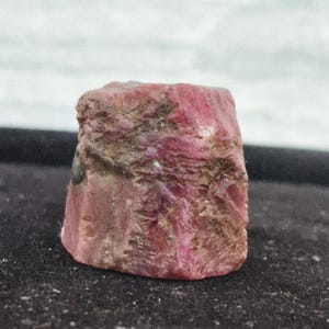 Könnte beinhalten: Ein roher, unpolierter Rubinkristall mit zylindrischer Form und einem tiefen rötlich-pinken Farbton. Die Oberfläche des Edelsteins weist ein raues, strukturiertes Aussehen mit natürlichen mineralischen Einschlüssen und Farbvariationen auf.