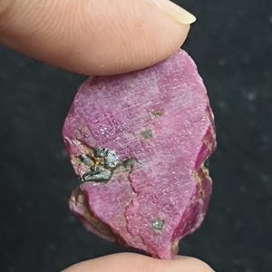 May include: A raw, unpolished ruby gemstone with a deep magenta-pink hue. The stone features a rough, textured surface with visible striations and small, dark metallic inclusions embedded within the crystalline structure of the mineral specimen.