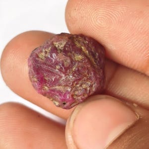 May include: A raw, unpolished ruby gemstone with a deep red colour and irregular, textured surface featuring patches of brownish-gold mineral inclusions. The stone has a multifaceted, natural shape with visible striations and a matte finish.