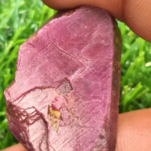 Könnte beinhalten: Ein roher, unpolierter Rubin-Edelstein mit einem tiefen Magenta- und Rosaton. Der Stein weist eine natürliche, unregelmäßige Form mit sichtbaren Oberflächentexturen, inneren Brüchen und kristallinen Facetten im gesamten Körper auf.